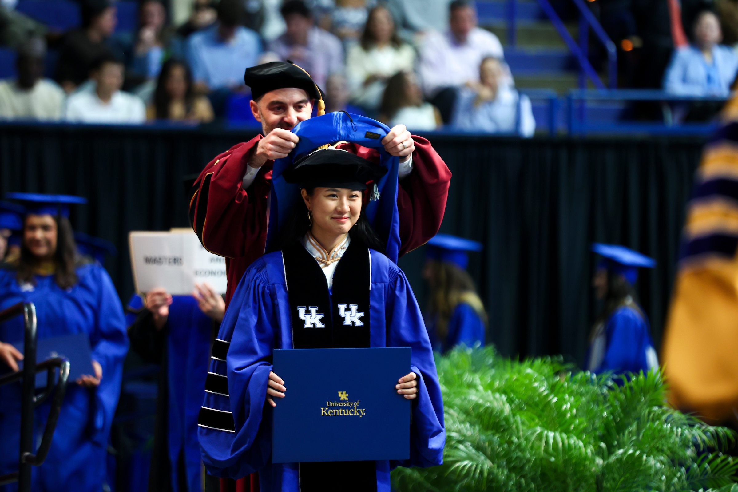 a doctoral student receives her hood at the commencement ceremony. Other graduates are blurry in the background 