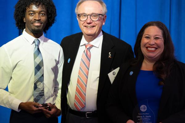 2025 Gatton College Lyman T. Johnson Award Recipients Tayshawn Juantez Gooch and Olivia N Davis stand with a representative from the Alumni Association with their awards at the 2025 Lyman T. Johnson Awards ceremony. They are standing in front of a blue curtain backdrop, they are holding their awards which are made of glass and engraved with their names