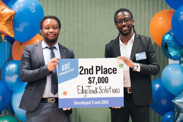 Two VACE participants stand with a sign between an orange and blue balloon arch. Both individuals are African American Males wearing dark colored suits. The sign they are holding says "GSE Collegiate Pitch, 2nd place, $7,000, EdgeTrack Solutions, Developed Track 2026." 