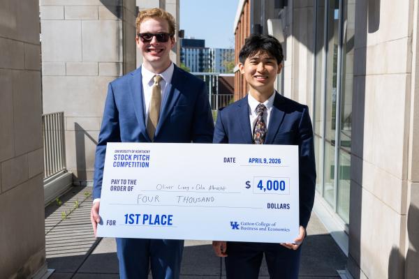Two people stand outside a building, holding a large check indicating a $4,000 prize. The check is awarded for 1st place in a stock pitch competition at the University of Kentucky, dated April 9, 2026. One individual is wearing sunglasses, and both are dressed in formal suits.