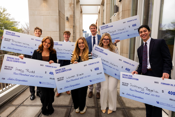 A large group of students, each holding a big check with their name on it, stand on an outdoor balcony space. All the students are smiling and celebratory and dressed professionally