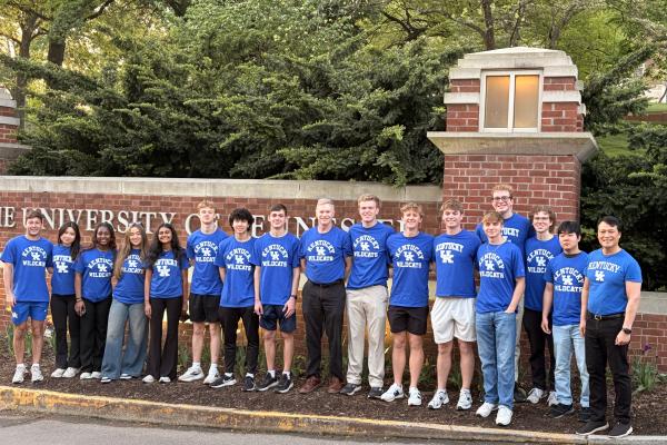 The Microsoft Excel Collegiate Challenge team is standing in front of a brick sign that reads "University of Tennessee." They are wearing matching blue "Kentucky Wildcats" shirts. Trees and greenery are visible in the background.