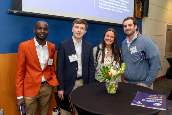 Four individuals are standing together around a small table at a professional event. The table holds a vase with flowers and a booklet. They appear to be smiling and dressed in business attire. A large screen in the background displays information, including the names UPS, DHL, Amazon, Intel, Toyota, and Walmart Global.