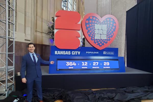 Daniel Wilmot stands in a suit next to a large display featuring the "Kansas City," "Populous," and "Purina" logos. The display includes the numbers 2026 and a heart with the words "We Are 26" on it. Below are countdown numbers showing 364 days, 12 hours, 27 minutes, and 29 seconds.