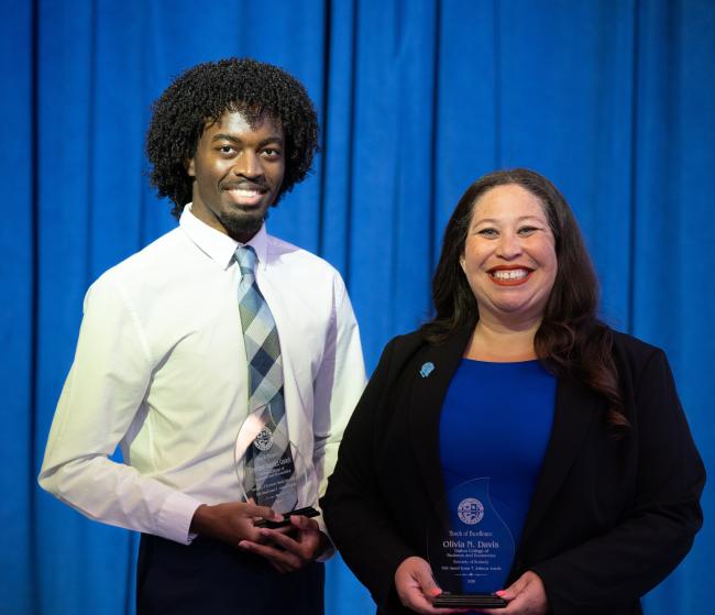 Award recipients Tayshawn Gooch and Olivia Davis stand with their glass awards in front of a blue curtain backdrop. Tayshawn is wearing a white collared shirt with a plaid tie while Olivia is wearing a kentucky blue dress with a black blazer. The glass awards have their names engraved on them.
