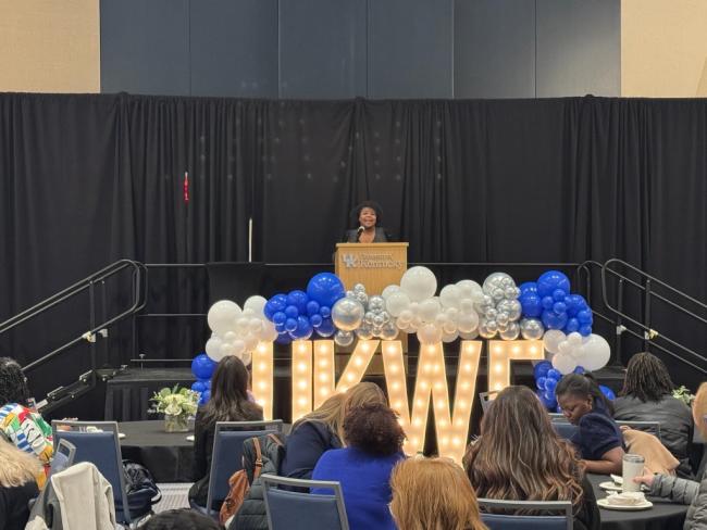 Ryessia Russell, director of Gatton Honors Pathways, speaks in front of the crowd at the annual Wildcat Forum. She stands behind a podium with Marquee letters in front spelling out "UKWF" and surrounded by blue and white balloons.