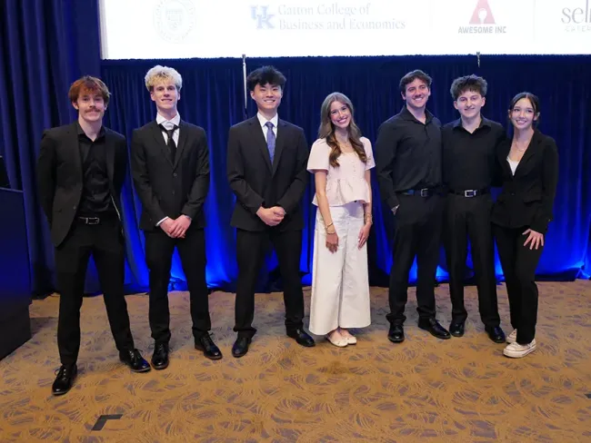 7 students pose for a picture in the front of Kincaid Auditorium. 6 of the students are wearing black outfits, while one is wearing white. They are all smiling.