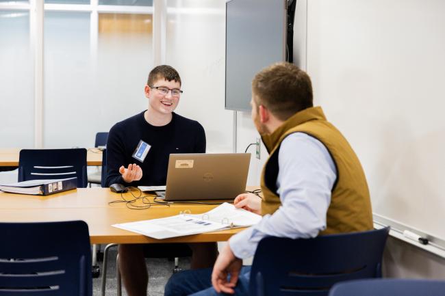 Two people are sitting at a table in a room. One person is using a Dell laptop, while the other has a notepad open. They appear to be having a conversation. The room has a whiteboard and a wall-mounted screen.