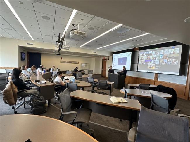 A classroom with students seated at desks and a presenter at the front; a large screen shows participants in a video conference call.