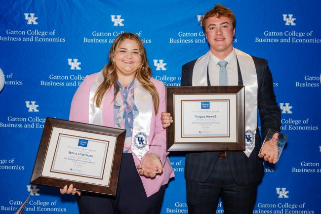 The two Dean's Award for Excellence Winners pose for a photo in front of a blue backdrop with the Gatton College of Business logo. Jenna, left, is wearing a pink blazer with a floral shirt. Teague, right, is wearing a dark suit with a white shirt and blue tie. Both students are holding a frame with a certificate, a glass award, and are wearing graduation stoles that say "Dean's Award for Excellence"