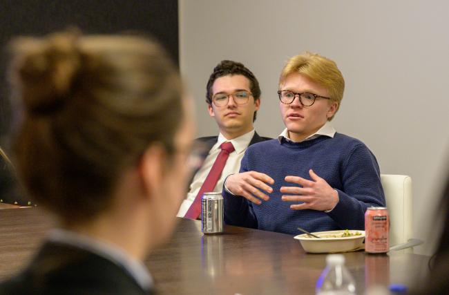A group of people sitting around a table in a meeting room. One individual is speaking and gesturing with hands, with a beverage can and a food container in front. Another person is observing attentively. Two drink cans are visible, one of which is a LaCroix can. The setting appears to be professional.