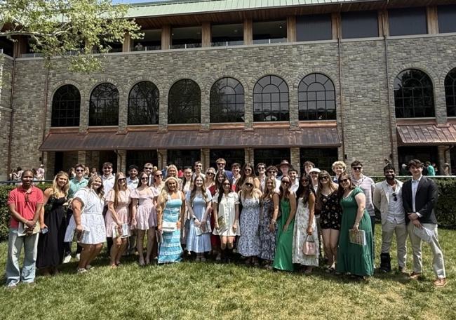 A group of people stands on a grassy area in front of a large brick building with arched windows. The scene is sunny, and the group appears dressed for a summer event, with a mix of casual and semi‑formal attire.