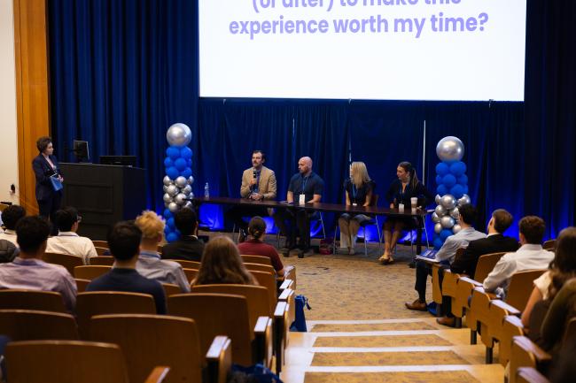 Audience listening to a panel discussion in a lecture hall. Four individuals seated at a table on stage with a large screen behind them displaying partially visible text. Blue and silver balloon decorations are on the stage.