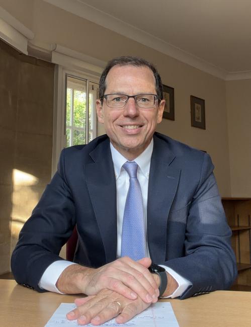 Carlos Lamarche wearing a business suit sitting at a desk, smiling at the camera, with papers and a watch visible on his wrist.