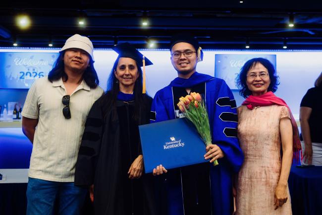 A group of four people stands together at a graduation event. One person is wearing a cap and gown, holding a University of Kentucky diploma and a bouquet of tulips. The background features a graduation sign.