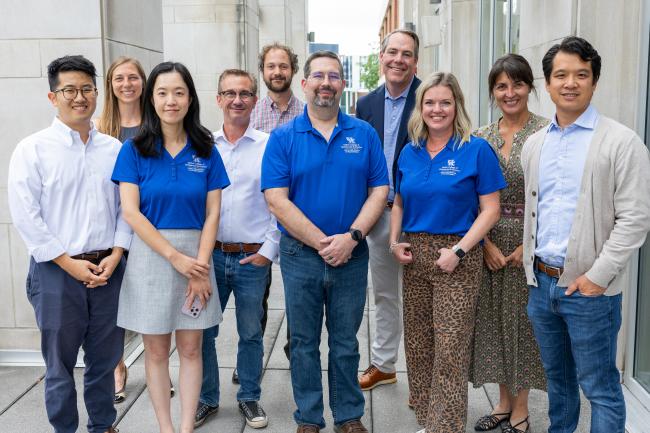 A group of individuals stands together outdoors in front of a building. Several people are wearing matching blue shirts with a logo that includes the Gatton College of Business and Economics logo. The group is smiling and appears to be posing for the photo.