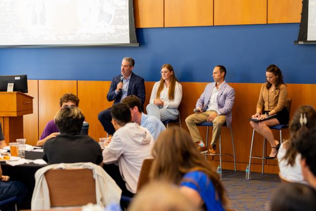 A panel of four individuals sits on stools at the front of a room during a conference or seminar. One person holds a microphone and speaks to an audience. Attendees are seated and listening attentively. In the background, a presentation screen is partially visible. The room has wood paneling and blue walls.