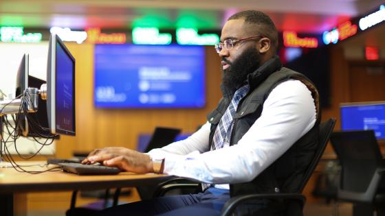 an African American graduate student sits at a computer in the finance center at Gatton as the stock ticker is blurred in the background