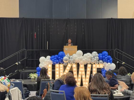 Ryessia Russell, director of Gatton Honors Pathways, speaks in front of the crowd at the annual Wildcat Forum. She stands behind a podium with Marquee letters in front spelling out "UKWF" and surrounded by blue and white balloons.