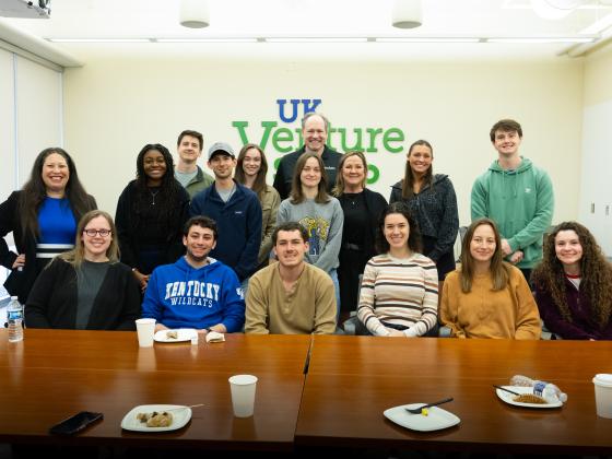 A group of people gathered in a room with a "UK Venture Studio" sign on the wall. They are seated and standing around a long wooden table with cups, a juice bottle, and some snacks visible. The setting appears to be casual and social.