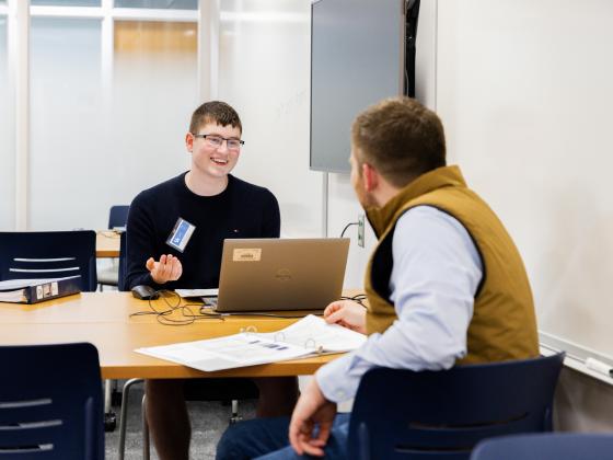 Two people are sitting at a table in a room. One person is using a Dell laptop, while the other has a notepad open. They appear to be having a conversation. The room has a whiteboard and a wall-mounted screen.