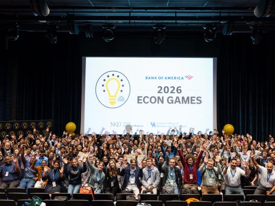 A large group of students stands together in an auditorium setting and poses for a picture. They are all excited, smiling, and raising their hands in the air in celebration. A large screen behind them reads "Bank of America 2026 Econ Games" with the Econ Games logo next to it- a yellow light bulb in a black circle.