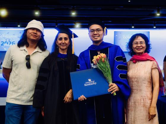 A group of four people stands together at a graduation event. One person is wearing a cap and gown, holding a University of Kentucky diploma and a bouquet of tulips. The background features a graduation sign.