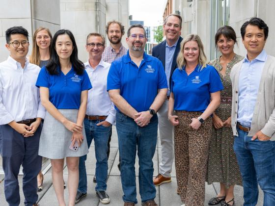 A group of individuals stands together outdoors in front of a building. Several people are wearing matching blue shirts with a logo that includes the Gatton College of Business and Economics logo. The group is smiling and appears to be posing for the photo.