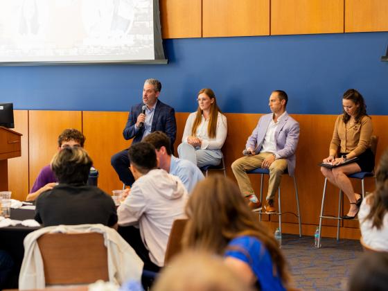A panel of four individuals sits on stools at the front of a room during a conference or seminar. One person holds a microphone and speaks to an audience. Attendees are seated and listening attentively. In the background, a presentation screen is partially visible. The room has wood paneling and blue walls.