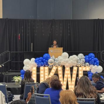 Ryessia Russell, director of Gatton Honors Pathways, speaks in front of the crowd at the annual Wildcat Forum. She stands behind a podium with Marquee letters in front spelling out "UKWF" and surrounded by blue and white balloons.