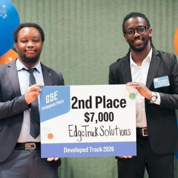 Two VACE participants stand with a sign between an orange and blue balloon arch. Both individuals are African American Males wearing dark colored suits. The sign they are holding says "GSE Collegiate Pitch, 2nd place, $7,000, EdgeTrack Solutions, Developed Track 2026." 