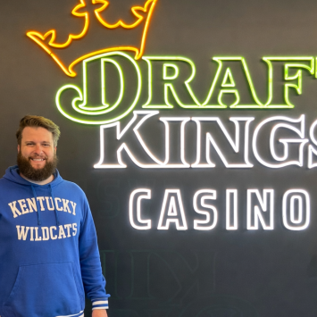 Brian Scroggins, a Gatton alum, poses for a picture in front of a neon sign that reads "Draft Kings Casino." He is wearing a UK blue sweatshirt that reads "Kentucky Wildcats."