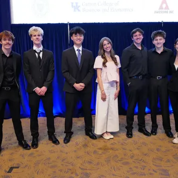 7 students pose for a picture in the front of Kincaid Auditorium. 6 of the students are wearing black outfits, while one is wearing white. They are all smiling.