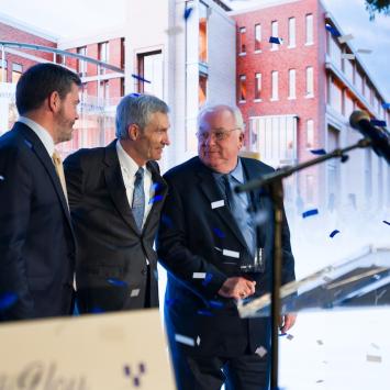 Three male individuals stand in front of a screen depicting a large building. They are looking at each other and smiling, one is holding a glass of wine. There's a podium in the foreground with a logo that reads "Gatton College of Business and Economics 100th year anniversary." There is confetti streaming down in the foreground as well.