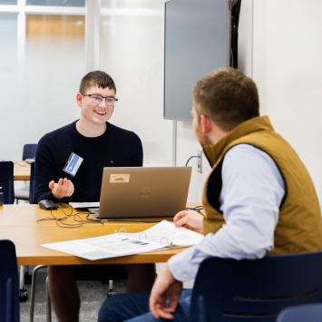 Two people are sitting at a table in a room. One person is using a Dell laptop, while the other has a notepad open. They appear to be having a conversation. The room has a whiteboard and a wall-mounted screen.