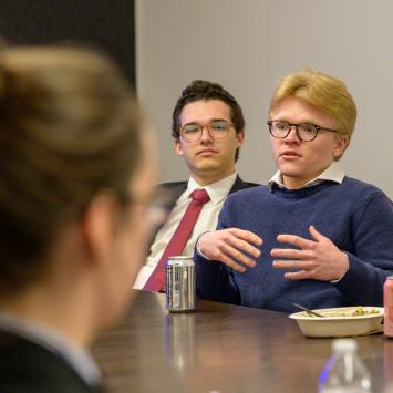 A group of people sitting around a table in a meeting room. One individual is speaking and gesturing with hands, with a beverage can and a food container in front. Another person is observing attentively. Two drink cans are visible, one of which is a LaCroix can. The setting appears to be professional.