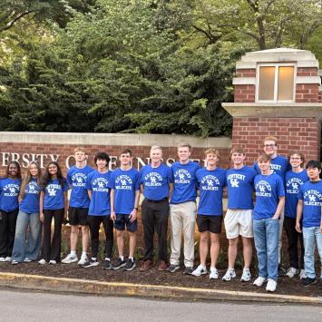 The Microsoft Excel Collegiate Challenge team is standing in front of a brick sign that reads "University of Tennessee." They are wearing matching blue "Kentucky Wildcats" shirts. Trees and greenery are visible in the background.