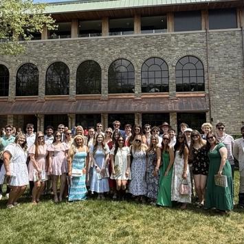 A group of people stands on a grassy area in front of a large brick building with arched windows. The scene is sunny, and the group appears dressed for a summer event, with a mix of casual and semi‑formal attire.