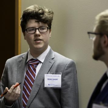 two individuals in formal attire, engaged in a conversation. One is holding a pen and wearing a name tag, standing in a room with beige walls.
