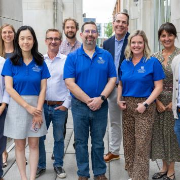 A group of individuals stands together outdoors in front of a building. Several people are wearing matching blue shirts with a logo that includes the Gatton College of Business and Economics logo. The group is smiling and appears to be posing for the photo.
