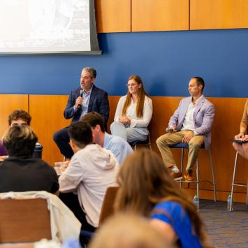 A panel of four individuals sits on stools at the front of a room during a conference or seminar. One person holds a microphone and speaks to an audience. Attendees are seated and listening attentively. In the background, a presentation screen is partially visible. The room has wood paneling and blue walls.